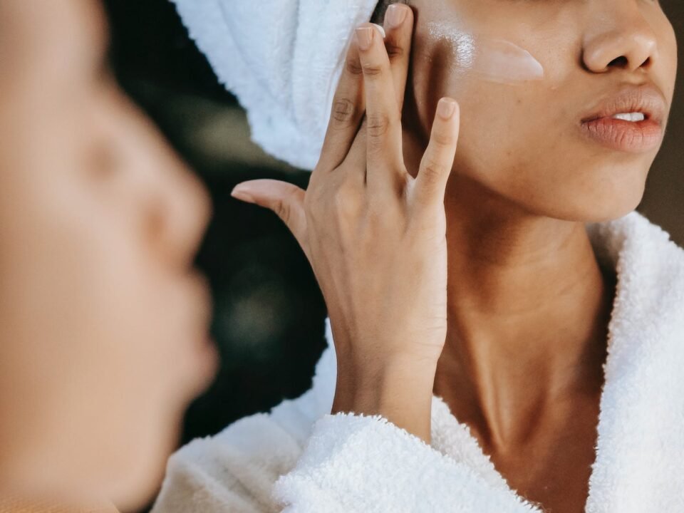 crop ethnic woman applying cream on face against mirror